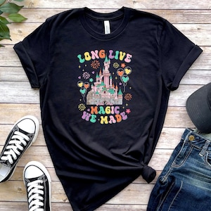 May include: A black short-sleeve t-shirt featuring a graphic of a castle with fireworks and balloons. The text reads LONG LIVE all the MAGIC WE MADE in colorful, retro-style lettering. The design includes pastel pink, blue, and yellow accents.