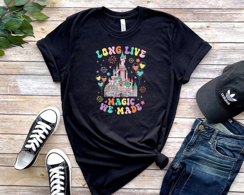 May include: A black short-sleeve t-shirt featuring a graphic of a castle with fireworks and balloons. The text reads LONG LIVE all the MAGIC WE MADE in colorful, retro-style lettering. The design includes pastel pink, blue, and yellow accents.