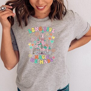 May include: A heather gray t-shirt featuring a colorful graphic of a castle with fireworks and balloons. The text reads LONG LIVE ALL THE MAGIC WE MADE in a multi-colored, rounded font. The design includes pastel pink, blue, and yellow accents.
