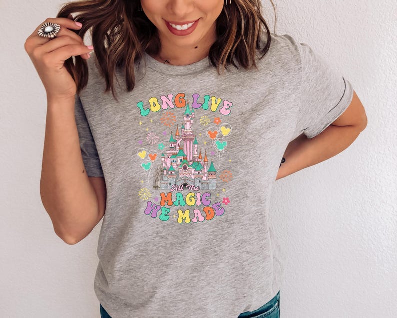 May include: A heather gray t-shirt featuring a colorful graphic of a castle with fireworks and balloons. The text reads LONG LIVE ALL THE MAGIC WE MADE in a multi-colored, rounded font. The design includes pastel pink, blue, and yellow accents.