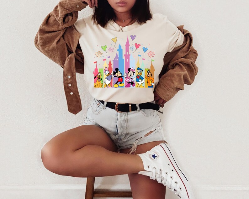 May include: A cream-colored t-shirt featuring a colorful graphic print of a castle silhouette and various cartoon characters including Mickey Mouse, Minnie Mouse, Donald Duck, Daisy Duck, Pluto, and Goofy, accented by floating balloons and fireworks.