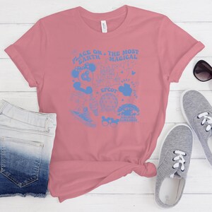 May include: A dusty rose short-sleeve t-shirt featuring a light blue graphic print. The design includes the text "PLACE ON THE EARTH THE MOST MAGICAL" along with various icons including a castle, a globe, and mouse ears, plus the word "EPCOT".