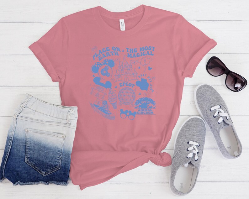 May include: A dusty rose short-sleeve t-shirt featuring a light blue graphic print. The design includes the text "PLACE ON THE EARTH THE MOST MAGICAL" along with various icons including a castle, a globe, and mouse ears, plus the word "EPCOT".