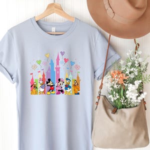 May include: A light blue short-sleeve t-shirt featuring a graphic print of Disney characters including Mickey Mouse, Minnie Mouse, Donald Duck, Daisy Duck, Pluto, and Goofy standing in front of a colorful castle with balloons and fireworks.