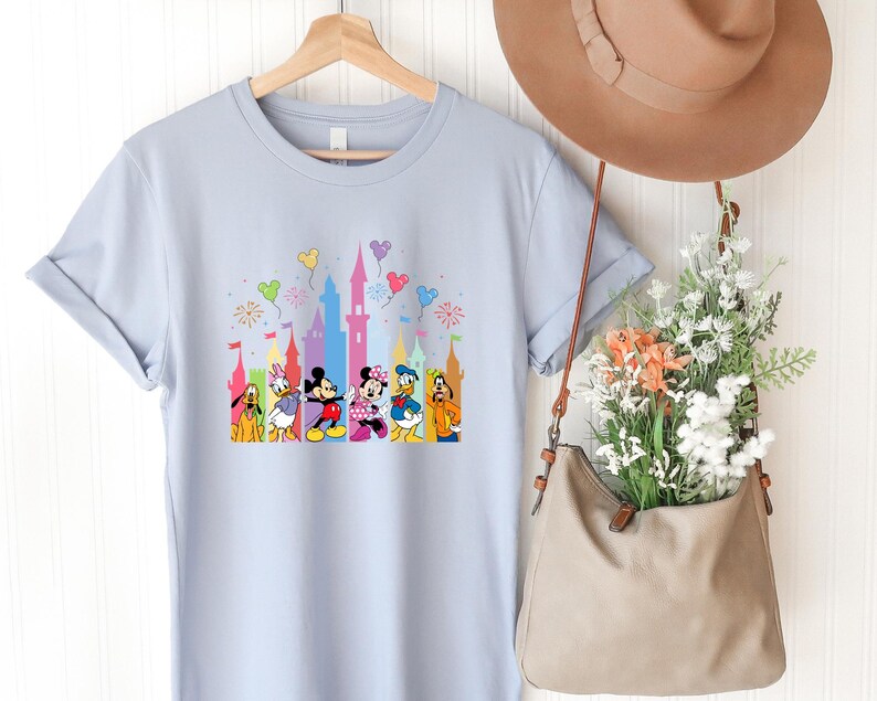 May include: A light blue short-sleeve t-shirt featuring a graphic print of Disney characters including Mickey Mouse, Minnie Mouse, Donald Duck, Daisy Duck, Pluto, and Goofy standing in front of a colorful castle with balloons and fireworks.