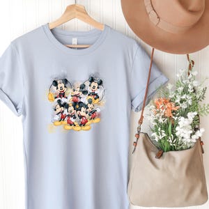 Op de afbeelding: Een lichtblauw t-shirt met korte mouwen en een centrale grafische print van meerdere Mickey Mouse-figuren in verschillende houdingen met gele en grijze accenten in aquarel-stijl. Het shirt hangt aan een houten kledinghanger.
