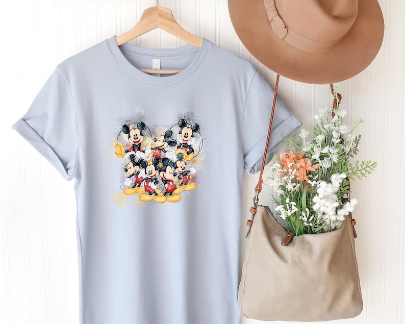 Op de afbeelding: Een lichtblauw t-shirt met korte mouwen en een centrale grafische print van meerdere Mickey Mouse-figuren in verschillende houdingen met gele en grijze accenten in aquarel-stijl. Het shirt hangt aan een houten kledinghanger.