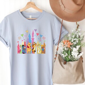May include: A light blue t-shirt featuring a graphic print of Winnie the Pooh characters including Piglet, Eeyore, Pooh, Tigger, Rabbit, and Roo standing in front of a colorful castle silhouette with floating balloons and fireworks above them.