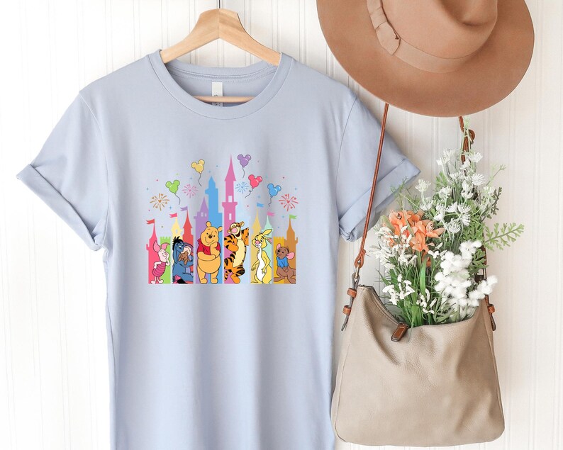 May include: A light blue t-shirt featuring a graphic print of Winnie the Pooh characters including Piglet, Eeyore, Pooh, Tigger, Rabbit, and Roo standing in front of a colorful castle silhouette with floating balloons and fireworks above them.