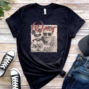 Puede incluir: Una camiseta negra de manga corta con un estampado gr&aacute;fico de Walt Disney y Mickey Mouse, ambos con gafas de sol. La palabra WALT est&aacute; impresa en una fuente roja estilizada sobre las figuras, sobre un fondo cuadrado de color claro.