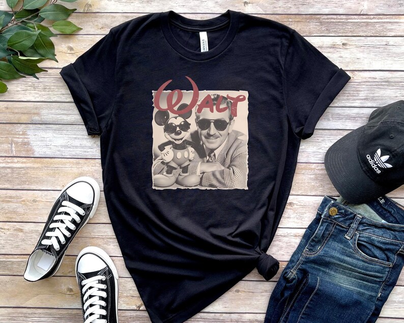 Puede incluir: Una camiseta negra de manga corta con un estampado gr&aacute;fico de Walt Disney y Mickey Mouse, ambos con gafas de sol. La palabra WALT est&aacute; impresa en una fuente roja estilizada sobre las figuras, sobre un fondo cuadrado de color claro.