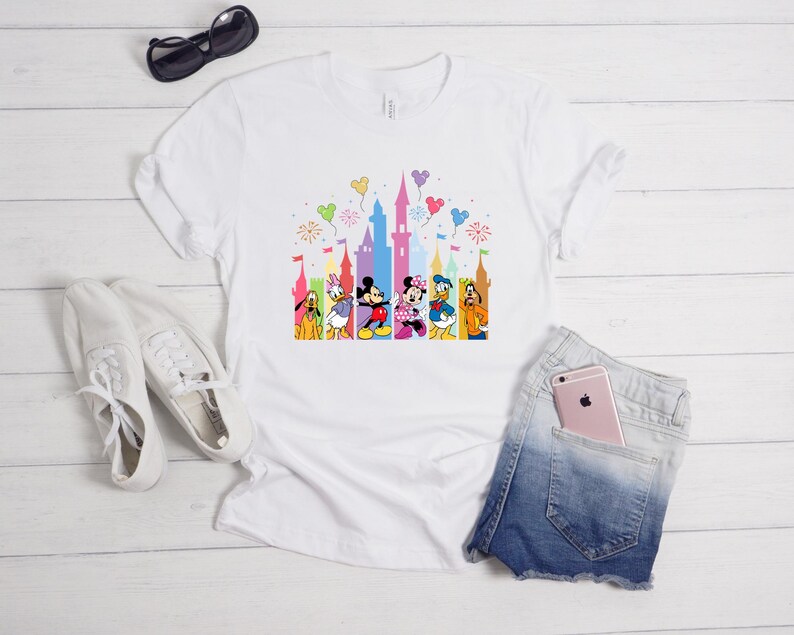 May include: A white short-sleeved t-shirt featuring a colorful graphic print of a castle silhouette and various cartoon characters including Mickey Mouse, Minnie Mouse, Donald Duck, Daisy Duck, Pluto, and Goofy, accented by floating balloons.