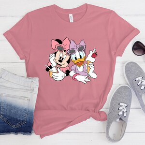 May include: A dusty rose short-sleeved t-shirt featuring a graphic print of Minnie Mouse and Daisy Duck wearing sunglasses and holding ice cream cones. The shirt is displayed alongside a pair of blue denim shorts and grey canvas trainers.
