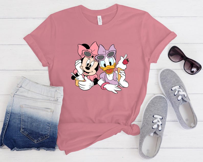May include: A dusty rose short-sleeved t-shirt featuring a graphic print of Minnie Mouse and Daisy Duck wearing sunglasses and holding ice cream cones. The shirt is displayed alongside a pair of blue denim shorts and grey canvas trainers.