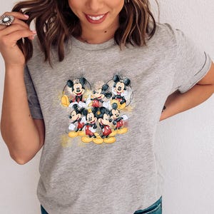 Op de afbeelding: Een grijs gem&ecirc;leerd T-shirt met korte mouwen en een centrale grafische print van meerdere Mickey Mouse-figuren in verschillende houdingen. Het ontwerp bevat gele en gouden aquarelachtige spetters rondom de zwarte, witte en rode figuren.
