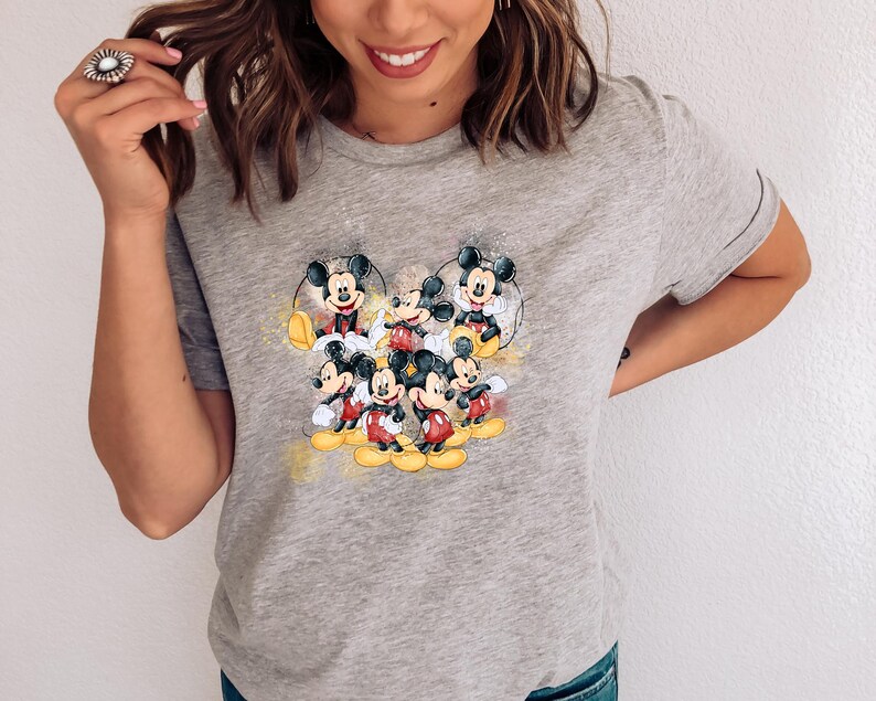 Op de afbeelding: Een grijs gem&ecirc;leerd T-shirt met korte mouwen en een centrale grafische print van meerdere Mickey Mouse-figuren in verschillende houdingen. Het ontwerp bevat gele en gouden aquarelachtige spetters rondom de zwarte, witte en rode figuren.