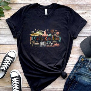 May include: A black short-sleeved t-shirt featuring a colourful graphic print in the centre with the text Magic Kingdom surrounded by various small illustrations of park attractions, buildings, and icons in shades of red, blue, green, and yellow.
