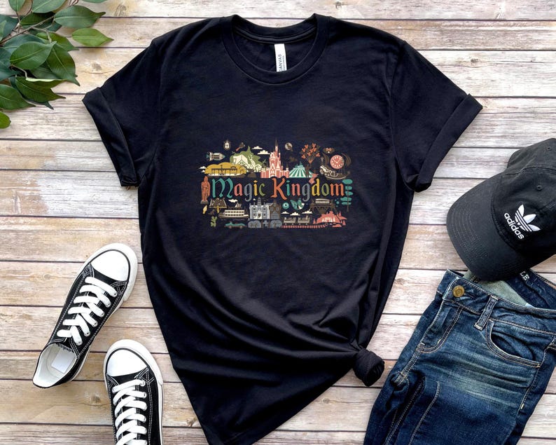 May include: A black short-sleeved t-shirt featuring a colourful graphic print in the centre with the text Magic Kingdom surrounded by various small illustrations of park attractions, buildings, and icons in shades of red, blue, green, and yellow.