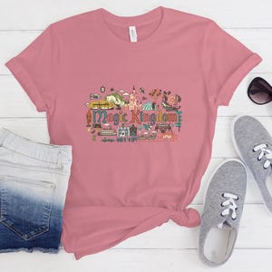 May include: A dusty rose short-sleeve t-shirt featuring a colourful graphic illustration of the Magic Kingdom theme park with various attractions and the text Magic Kingdom printed in the centre. The shirt is paired with denim shorts and trainers.
