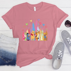 May include: A dusty rose short-sleeved t-shirt featuring a colorful graphic print of Winnie the Pooh characters including Piglet, Eeyore, Pooh, Tigger, Rabbit, and Roo standing in front of a castle silhouette with floating balloons above them.