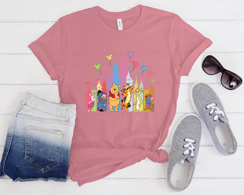 May include: A dusty rose short-sleeved t-shirt featuring a colorful graphic print of Winnie the Pooh characters including Piglet, Eeyore, Pooh, Tigger, Rabbit, and Roo standing in front of a castle silhouette with floating balloons above them.