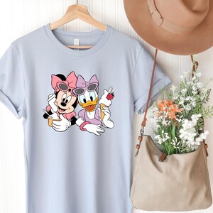May include: A light blue t-shirt featuring a graphic print of Minnie Mouse and Daisy Duck wearing pink bows and sunglasses while holding ice cream cones. The shirt is displayed on a wooden hanger next to a tan hat and a tan bag filled with flowers.