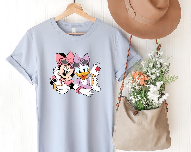 May include: A light blue t-shirt featuring a graphic print of Minnie Mouse and Daisy Duck wearing pink bows and sunglasses while holding ice cream cones. The shirt is displayed on a wooden hanger next to a tan hat and a tan bag filled with flowers.