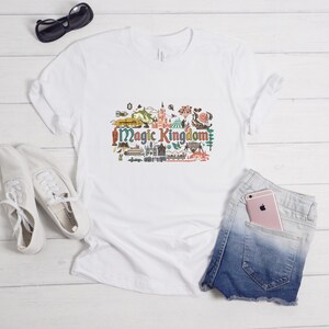 May include: A white short-sleeved t-shirt featuring a colourful graphic print of the text Magic Kingdom surrounded by various small illustrations of theme park landmarks, attractions, and icons in shades of red, green, blue, and yellow.