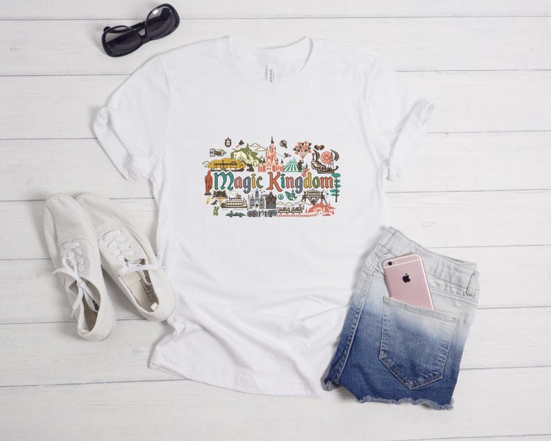May include: A white short-sleeved t-shirt featuring a colourful graphic print of the text Magic Kingdom surrounded by various small illustrations of theme park landmarks, attractions, and icons in shades of red, green, blue, and yellow.