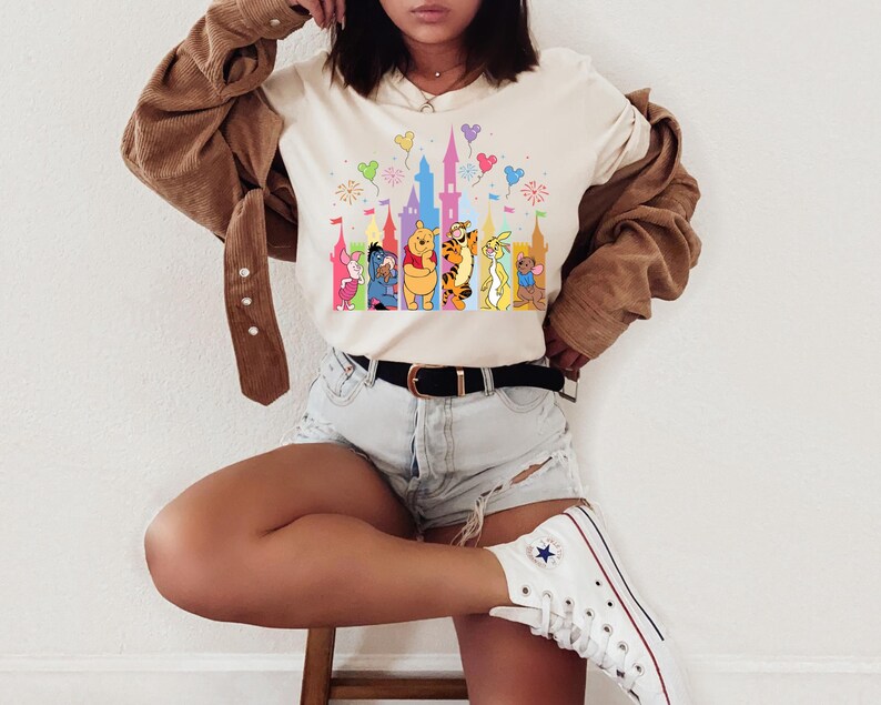 May include: A cream-colored short-sleeve t-shirt featuring a colorful graphic print of a castle silhouette with fireworks and balloons, accompanied by illustrations of Piglet, Eeyore, Winnie the Pooh, Tigger, Roo, and Rabbit standing in a row.