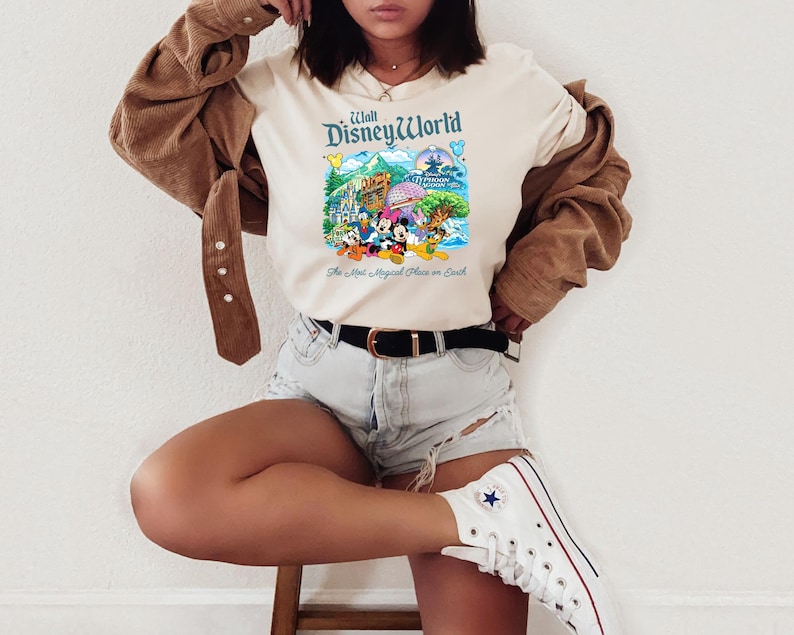 Op de afbeelding: Een cr&egrave;mekleurig t-shirt met een kleurrijke afbeelding van Walt Disney World met personages zoals Mickey Mouse, Minnie Mouse, Donald Duck en Pluto, en de tekst The Most Magical Place on Earth onder de illustratie gedrukt.