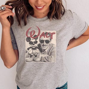 Puede incluir: Una camiseta gris jaspeado de manga corta con un estampado gr&aacute;fico de una fotograf&iacute;a en blanco y negro que muestra a un hombre y un rat&oacute;n de dibujos animados con gafas de sol. La palabra Walt est&aacute; impresa en una fuente roja grande.