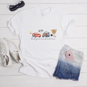 May include: A white short-sleeve t-shirt featuring a graphic of two cartoon cars, a trophy, checkered flags, and a sign that reads Cozy Cone Motel. Below the cars, the text reads Find your own finish line in a cursive black font.