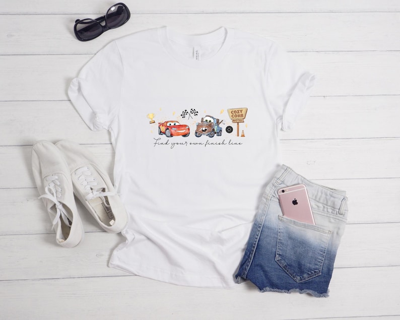 May include: A white short-sleeve t-shirt featuring a graphic of two cartoon cars, a trophy, checkered flags, and a sign that reads Cozy Cone Motel. Below the cars, the text reads Find your own finish line in a cursive black font.