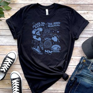 May include: A black short-sleeve t-shirt featuring a light blue graphic print with the text "PLACE ON THE MOST MAGICAL EARTH" and various theme park icons including a castle, a geodesic dome, mouse ears, sunglasses, and a trainer.