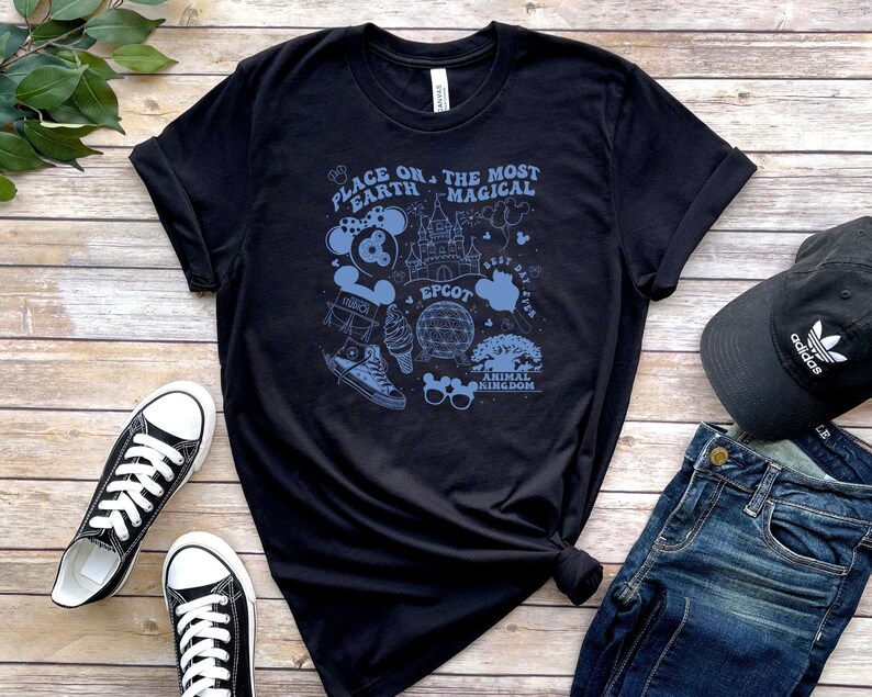 May include: A black short-sleeve t-shirt featuring a light blue graphic print with the text "PLACE ON THE MOST MAGICAL EARTH" and various theme park icons including a castle, a geodesic dome, mouse ears, sunglasses, and a trainer.