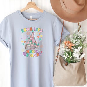May include: A light blue short-sleeve t-shirt featuring a graphic of a castle with the text "LONG LIVE" and "ALL THE MAGIC WE MADE" in colorful, retro-style lettering, surrounded by small hearts, flowers, and balloon icons in various pastel colors.