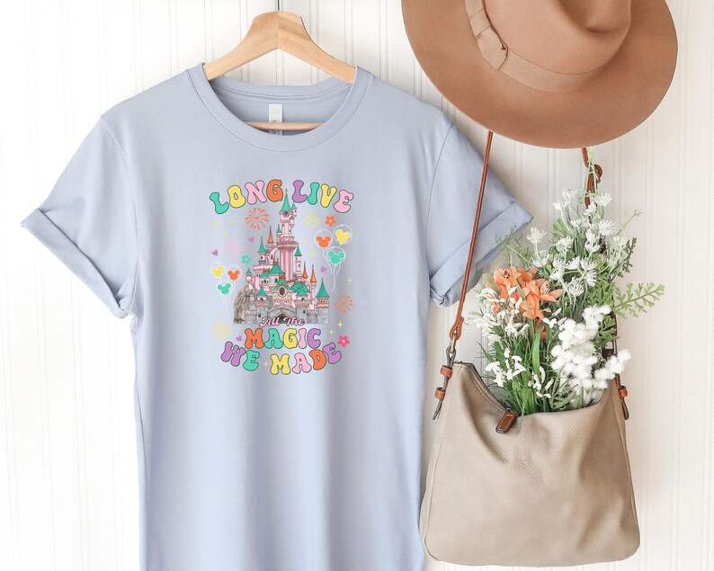 May include: A light blue short-sleeve t-shirt featuring a graphic of a castle with the text "LONG LIVE" and "ALL THE MAGIC WE MADE" in colorful, retro-style lettering, surrounded by small hearts, flowers, and balloon icons in various pastel colors.