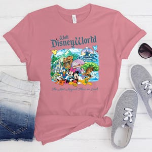 Op de afbeelding: Een oudroze T-shirt met korte mouwen en een kleurrijke afbeelding van Walt Disney World bezienswaardigheden en figuren, waaronder Mickey Mouse, Minnie Mouse, Donald Duck en Pluto, met de tekst "Walt Disney World" en "The Most Magical Place on Earth."