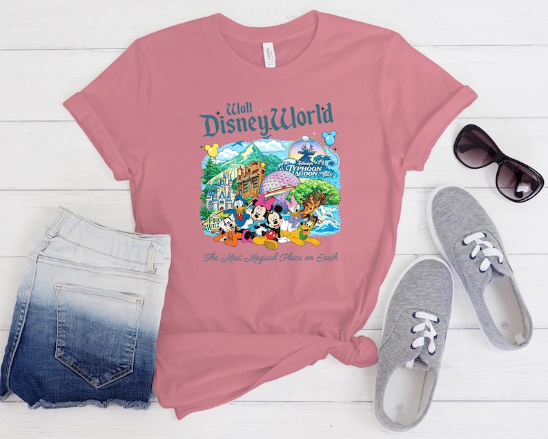 Op de afbeelding: Een oudroze T-shirt met korte mouwen en een kleurrijke afbeelding van Walt Disney World bezienswaardigheden en figuren, waaronder Mickey Mouse, Minnie Mouse, Donald Duck en Pluto, met de tekst "Walt Disney World" en "The Most Magical Place on Earth."
