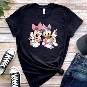 May include: A black short-sleeved t-shirt featuring a graphic print of Minnie Mouse and Daisy Duck wearing sunglasses and bows while eating ice cream. The characters are positioned in the centre of the shirt, which has a crew neckline and rolled sleeves.