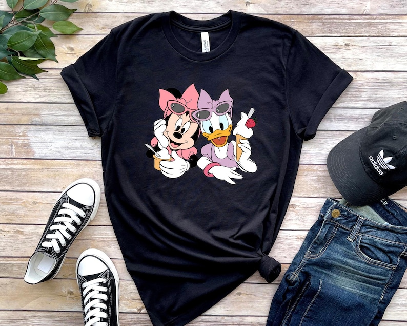 May include: A black short-sleeved t-shirt featuring a graphic print of Minnie Mouse and Daisy Duck wearing sunglasses and bows while eating ice cream. The characters are positioned in the centre of the shirt, which has a crew neckline and rolled sleeves.