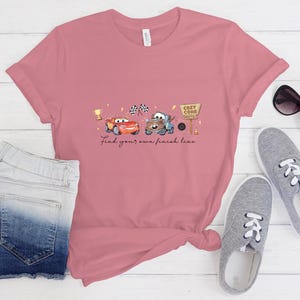 May include: A dusty rose short-sleeve t-shirt featuring a graphic of two cartoon cars, a gold trophy, checkered flags, and a Cozy Cone Hotel sign. The text below the graphic reads, Find your own finish line. The shirt is paired with denim shorts.
