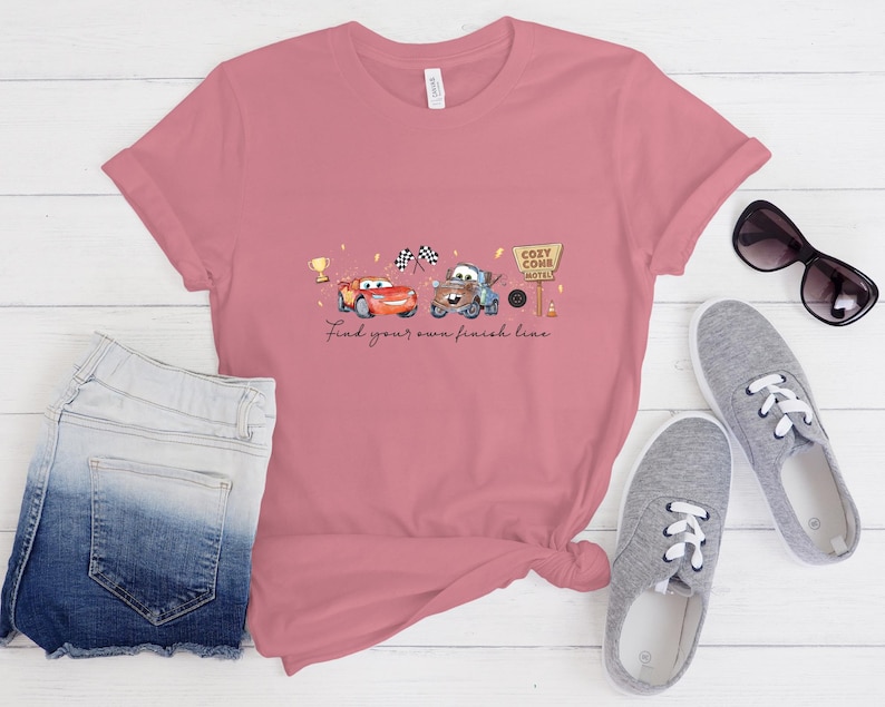 May include: A dusty rose short-sleeve t-shirt featuring a graphic of two cartoon cars, a gold trophy, checkered flags, and a Cozy Cone Hotel sign. The text below the graphic reads, Find your own finish line. The shirt is paired with denim shorts.