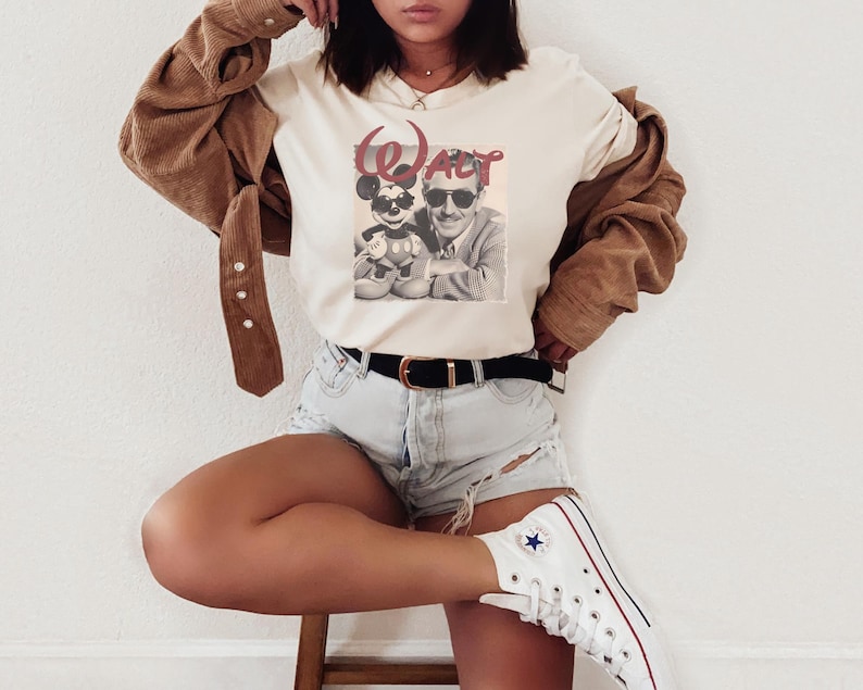 Puede incluir: Una camiseta de manga corta de color crema con un estampado gr&aacute;fico de una persona y un rat&oacute;n de dibujos animados, ambos con gafas de sol, y el texto rojo "WALT" arqueado encima. La prenda se combina con una camisa de pana marr&oacute;n, pantalones cortos de mezclilla azul claro, un cintur&oacute;n negro y zapatillas blancas de ca&ntilde;a alta.