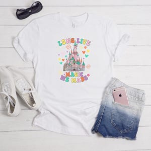 May include: A white short-sleeve t-shirt featuring a colorful graphic of a castle with the text "LONG LIVE THE MAGIC WE MADE" in a retro font, surrounded by small icons of fireworks, flowers, and mouse ears in pastel shades of pink, blue, and yellow.