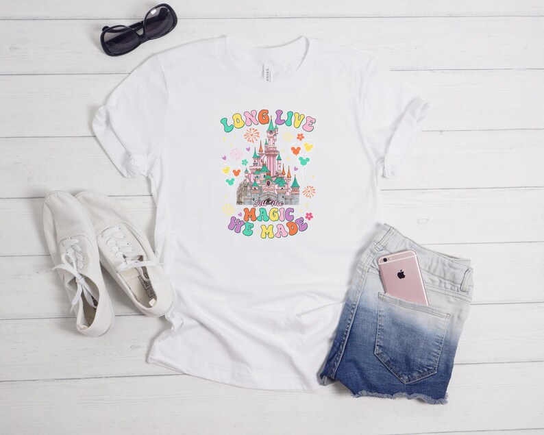 May include: A white short-sleeve t-shirt featuring a colorful graphic of a castle with the text "LONG LIVE THE MAGIC WE MADE" in a retro font, surrounded by small icons of fireworks, flowers, and mouse ears in pastel shades of pink, blue, and yellow.
