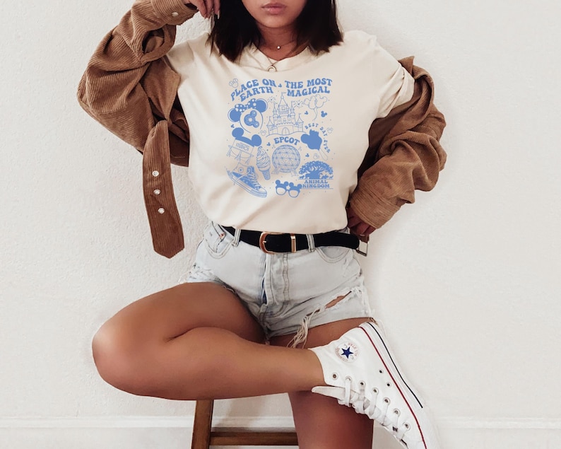 May include: A cream-coloured short-sleeve t-shirt featuring a blue graphic print with the text "Place on Earth the Most Magical" and "Epcot". The design includes illustrations of a castle, a globe, mouse ears, and other park-themed icons.