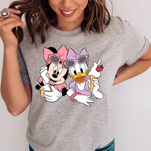 May include: A heather grey t-shirt featuring a graphic print of Minnie Mouse and Daisy Duck. Minnie wears a pink bow and sunglasses, holding a yellow cup. Daisy wears a purple bow and sunglasses, holding an ice cream cone with a strawberry.