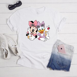 May include: A white short-sleeved t-shirt featuring a graphic print of Minnie Mouse and Daisy Duck wearing sunglasses and bows. Minnie holds a yellow cup with a spoon, while Daisy holds an ice cream cone topped with a strawberry.